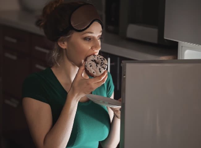 woman eating a cookie at night by refrigerator door