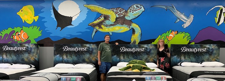 Artist John Alvey and Alithea Lass of William Thomas Mattress Gallery — holding store mascot Arlo the mini Pomeranian — pose in front of a mural honoring the new Beautyrest Harmony Lux collection with Seaqual ticking.